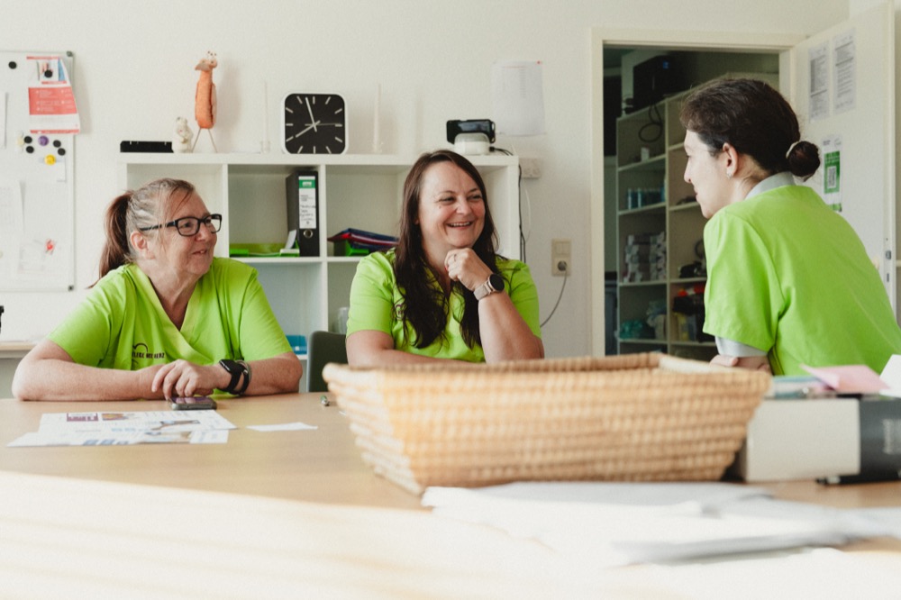 Drei Mitarbeiterinnen in grünen Shirts sitzen lachend am Tisch im Dienstzimmer und besprechen sich