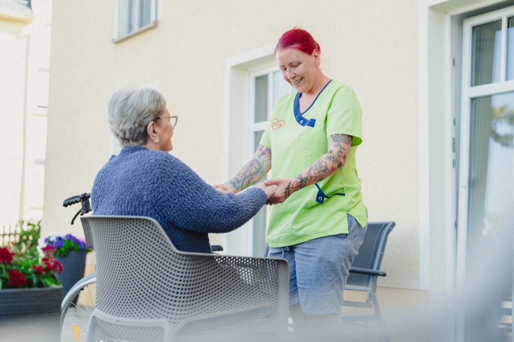 Eine Pflegekraft im grünen Kasack begrüßt eine Bewohnerin im Rollstuhl auf der Terrasse und hält ihre Hände.