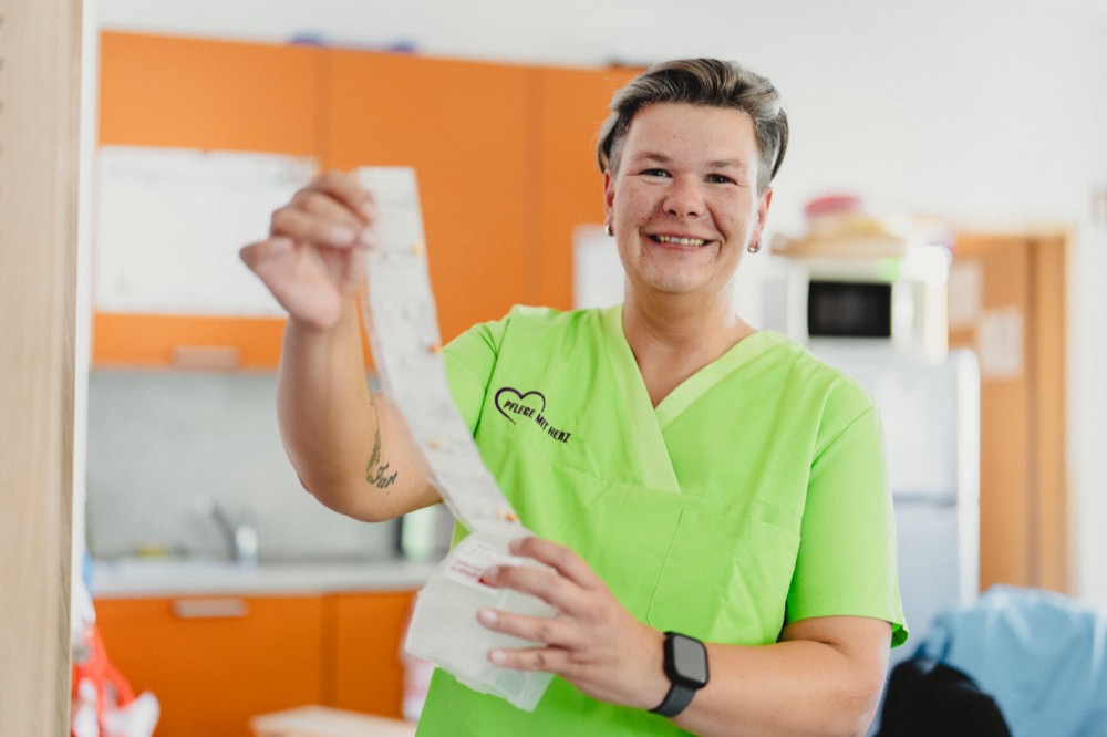 Pflegekraft in grüner Dienstkleidung lächelt in die Kamera und hält eine vorbereitete Medikamentenkarte in der Hand