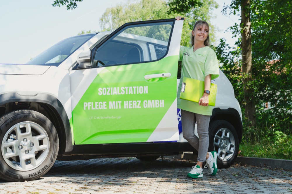 Pflegekraft in grüner Dienstkleidung steht mit einer Mappe an der offenen Tür des beschrifteten Dienstwagens
